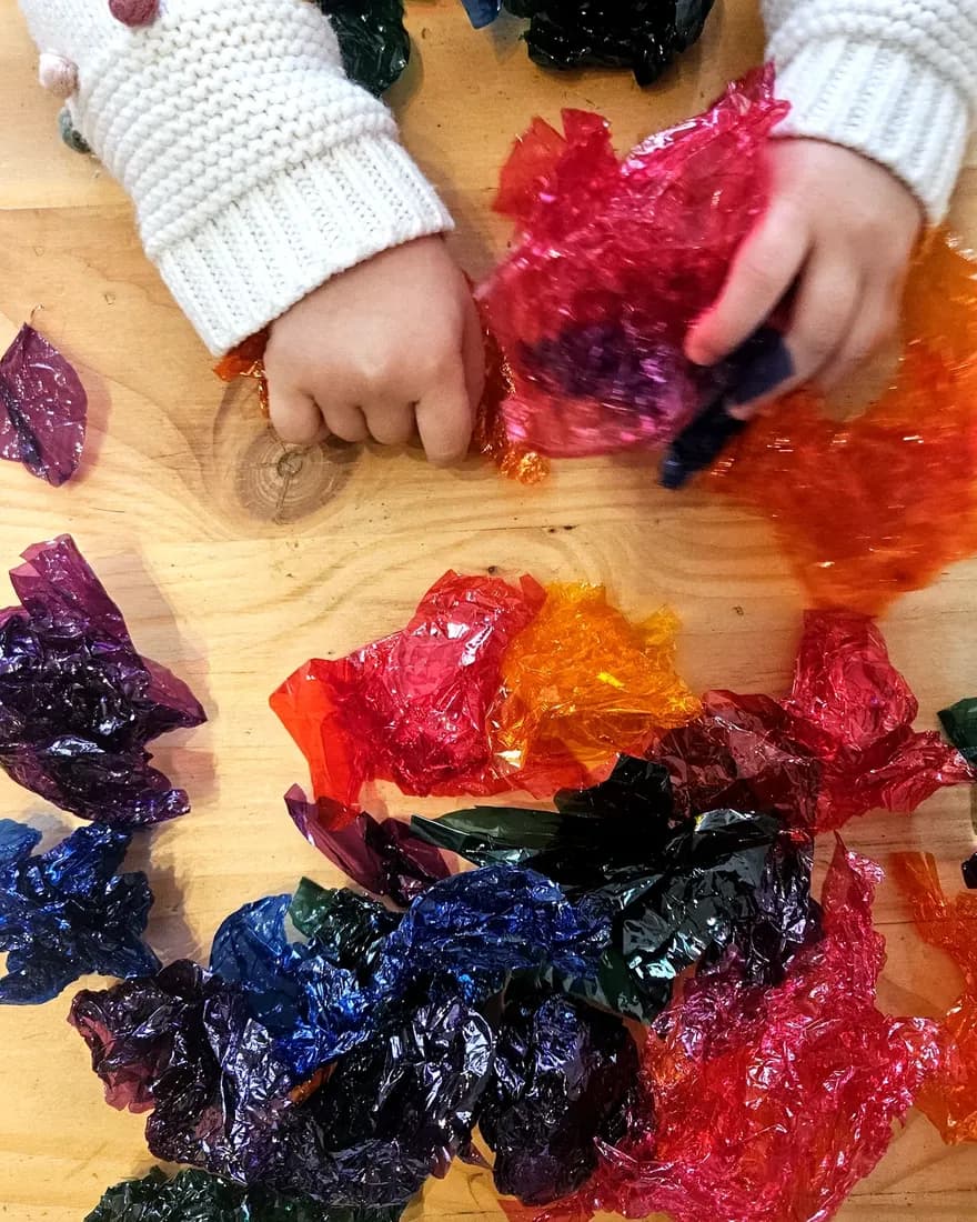 Manos de un niño jugando con papel celofán de colores sobre una mesa de madera.